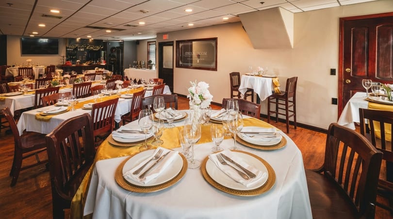 A restaurant dining room with tables set with white tablecloths, gold runners, and place settings.
