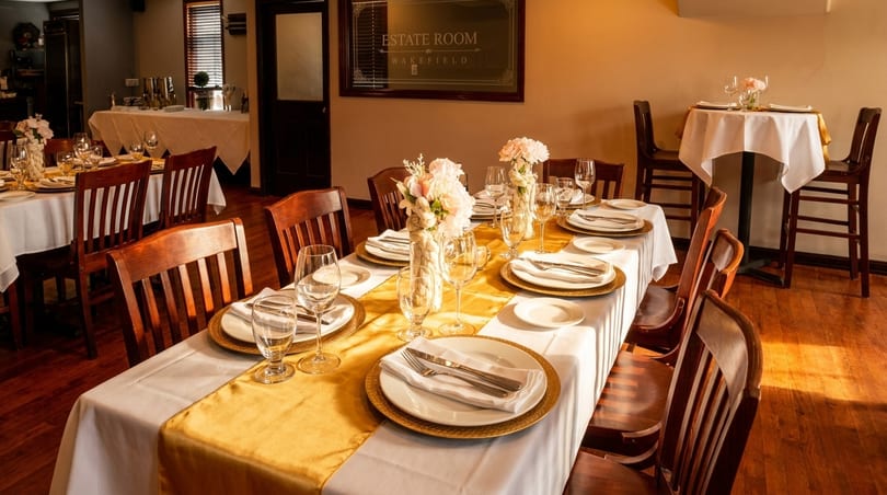 Elegant dining room with set tables, white tablecloths, gold runners, and floral centerpieces.