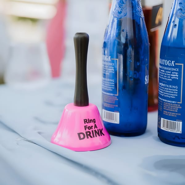 Pink "Ring For A DRINK" bell next to two blue Saratoga sparkling water bottles on a light blue tablecloth.