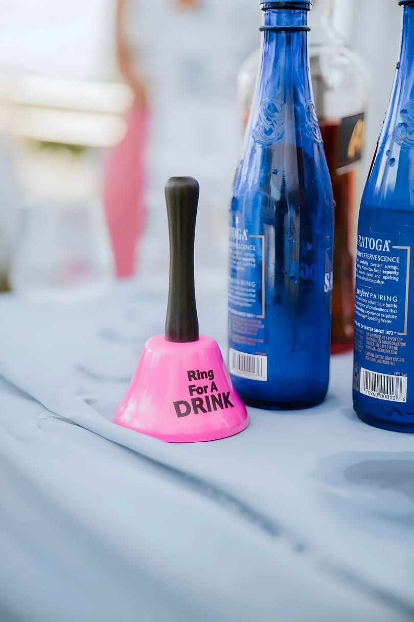 Pink "Ring For A DRINK" bell next to two blue Saratoga sparkling water bottles on a light blue tablecloth.