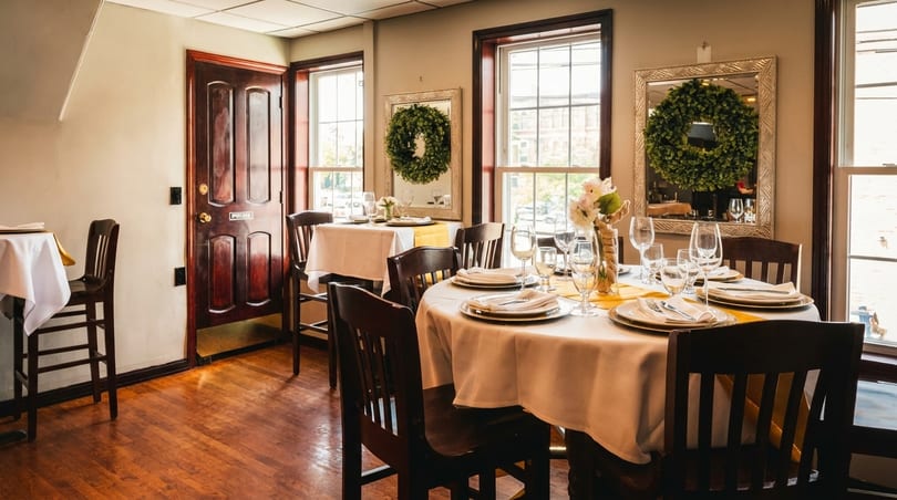 Restaurant dining room with set tables, dark wood chairs, and windows with wreaths reflecting in mirrors.