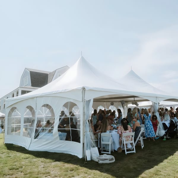 Guests seated at tables inside a large white tent on a grassy lawn next to a building.