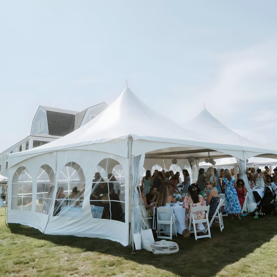 Guests seated at tables inside a large white tent on a grassy lawn next to a building.