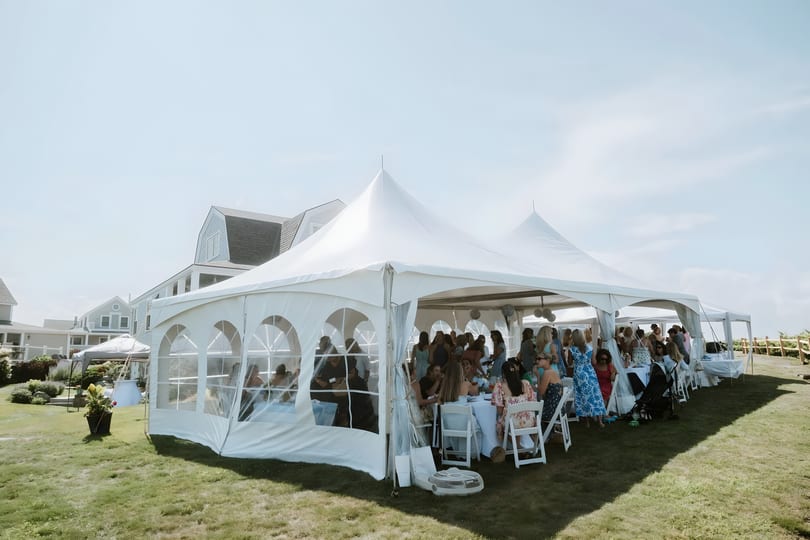 Guests seated at tables inside a large white tent on a grassy lawn next to a building.