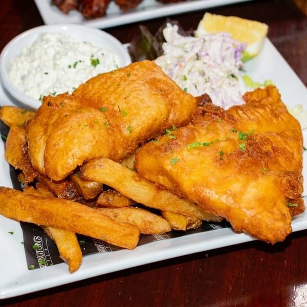 Golden-brown fish and chips with coleslaw, tartar sauce, and a lemon wedge on a white plate.