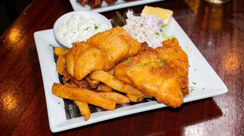 Golden-brown fish and chips with coleslaw, tartar sauce, and a lemon wedge on a white plate.