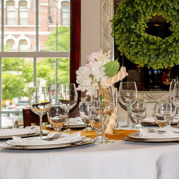 A dining table set with white tablecloth, wine glasses, and silverware, next to a window.