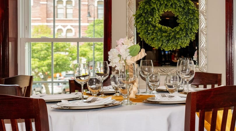 A dining table set with white tablecloth, wine glasses, and silverware, next to a window.
