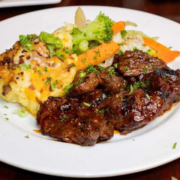 Grilled steak pieces with mashed potatoes, melted cheese, and mixed vegetables on a white plate.