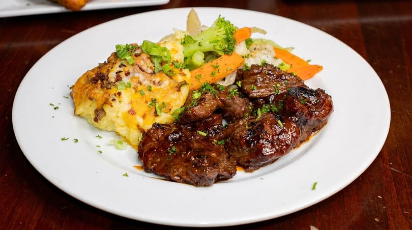 Grilled steak pieces with mashed potatoes, melted cheese, and mixed vegetables on a white plate.